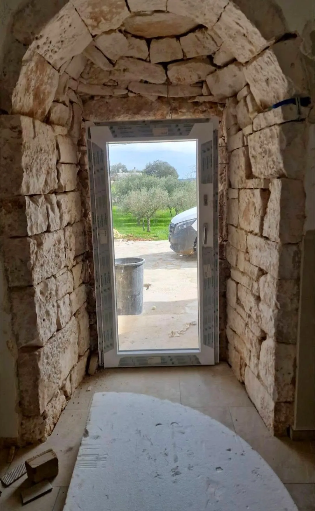 Portafinestra in PVC bianco montata sotto l'arco in pietra di un trullo, con vista sugli ulivi